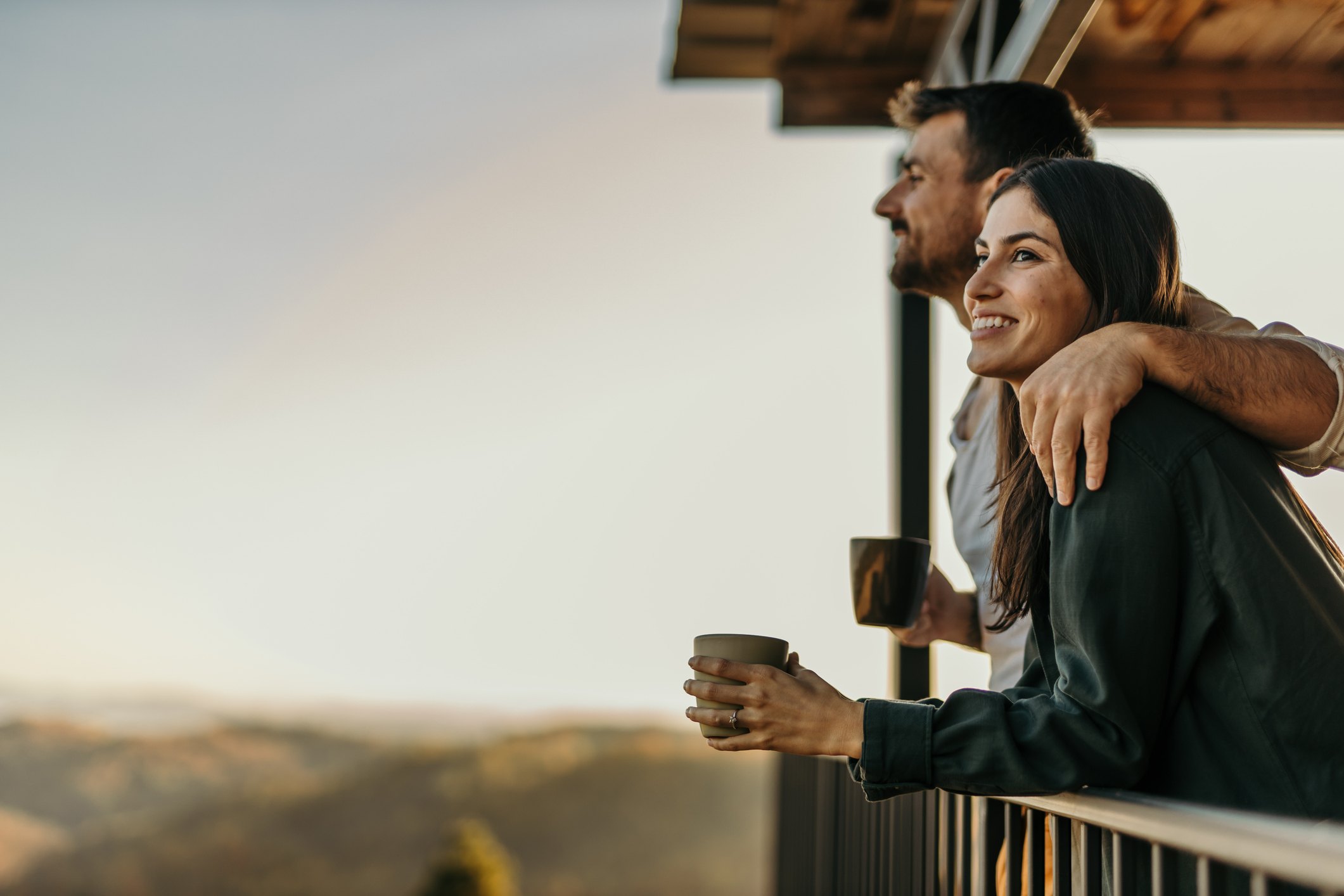 A couple holding coffee mugs on a balcony at sunset, looking out over rolling hills.
